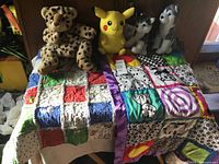 Two patchwork baby quilts laid side by side with four animal plush toys arranged on top and an animal backpack partially visible behind.