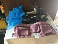 Photo showing a pink tool belt with several pockets, a black and white patterned baby carrier with straps, and a blue mesh laundry bag.