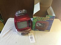 Curtis red retro TV with black & white screen, radio tuner visible on the right side of the front panel, placed next to the original box with colorful label showing product image. Accessories wrapped in plastic on top of TV.