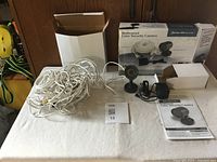 Full lot with long bundle of white cables, black power adapter, black security camera on mount, user manual, and packaging box.