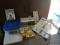 Photo showing Calmaroo Puppy toy box, educational games, selfie stick box, plastic containers and white drying rack arranged on a table near wooden cabinet with light coming from window.