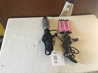 Two styling devices placed on a white surface, black round brush dryer and pink triple barrel curling iron with their cords wrapped around the handles.