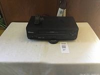 Full front view of black Memorex VCR on white table, remote control placed on top.