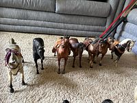 Front row of standing vintage Breyer model horses including a white horse with purple bridle, black horse, and brown horses with saddles arranged on carpet