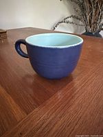 Blue ceramic cup with handle and light blue interior placed on a wooden table with decorative dried plants in background