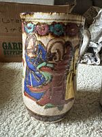 Hand-painted ceramic pitcher showing detailed multicolor figures and floral border