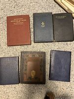 Six antique books laid out on carpet showing front covers and spines, different colors and embossed designs, with clear titles on some such as Merchant of Venice and The Man Without a Country.