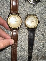 Two gold-tone watches: one LeCoultre-style guilloche dial with brown leather strap and one Gruen Precision with black strap