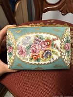 Photo of the top of the small vintage porcelain jewelry box with colorful hand-painted floral design surrounded by gold leaf accents and turquoise background.