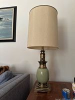 Full view of the vintage table lamp showing beige fabric shade and green ceramic brass base on a square brass metal platform stand near couch.