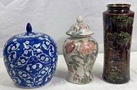 Three ceramic pieces grouped: blue and white covered jar, floral covered jar, and tall dark reddish brown vase with floral and bird motif