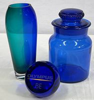 Grouping of all three cobalt glass items arranged together on a white background showing apothecary jar, paperweight, and bud vase