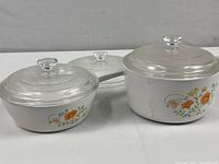 Image showing two white CorningWare covered sauce pans with Wildflower pattern and clear glass lids, one with a long handle and one with a side loop handle, plus an extra glass lid behind them.
