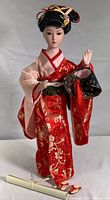 Full view of the porcelain Geisha doll standing, showing red and gold kimono, and scroll accessory on the floor in front