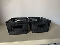 Two black plastic rectangular storage baskets with open grid woven pattern and built-in handles shown side by side on a flat surface.