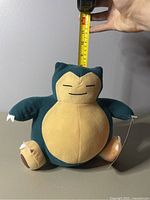 Front view of 8-inch Snorlax plush with hand and tape measure showing approximate height.