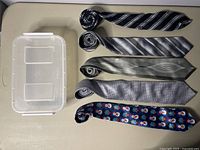Five assorted men's neckties laid flat showing different patterns and colors alongside a clear plastic storage container.