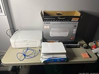 White Canon Pixma MG3620 printer with blue cables, stack of printer paper, Canon printer box, and a paper hole punch on table.