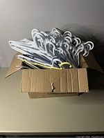 Box filled with approximately 100 plastic coat hangers in white, grey, and some yellow.