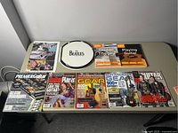 Books and magazines about guitar and music, along with a Beatles-branded drumhead, arranged on a table.