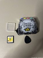 Guitar Hero DS game cartridge, Guitar Hero DS game accessory with fret buttons, and black guitar pick all displayed on flat surface.