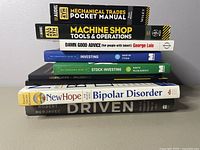Stack of 7 books with titles visible related to mechanics, investing, bipolar disorder, and personal development.