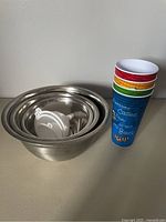 Photo showing 4 colorful M&M plastic drinking cups stacked and 3 nested stainless steel mixing bowls with 3 white plastic funnels inside the smallest bowl