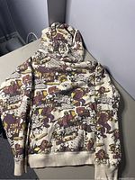 Full front view of the cream-colored sweater with multi-color lumberjack graffiti print laid flat on gray surface.