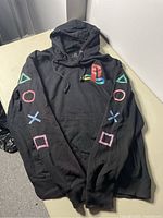 Front view of men’s large black PlayStation hoodie sweatshirt showing colorful embroidered PlayStation logo on chest and PlayStation controller symbols embroidered down both sleeves.