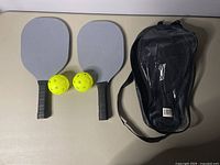Two gray pickleball paddles, two yellow pickleballs with holes, and black carrying bag on light surface
