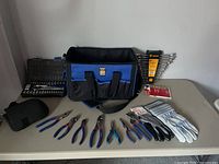 Full layout of tools on table including tool bag, socket set, wrenches, pliers, gloves, knee pads, and sanding paper