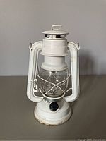 Left-side angled view of white metal lantern showing wear and rust at base and edges, with glass globe intact inside wire cage.