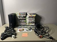 Front view showing black Xbox 360 console vertically with two controllers, power brick, AV and HDMI cables, four loose discs in front and stacks of game cases behind