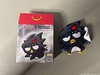 Front view of the plush with the red McDonalds box showing the Yu-Gi-Oh x Hello Kitty and Friends branding and a penguin character wearing a monster hoodie with red eyes.
