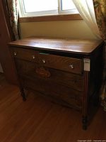 Full view of the antique wooden dresser in dark finish against a window with floral curtains, showing the four drawers and its condition.