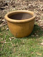 Brown ceramic urn positioned outdoors on grass with dry leaves around it, showing the urn's rounded mouth and exterior.