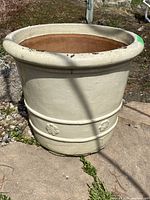 Image of a large cream-colored clay garden pot with decorative floral relief around the bottom band, showing cracks and paint chipping from outdoor use.