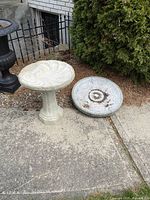Full view of assembled concrete bird bath next to separate cracked basin on concrete surface outdoors.