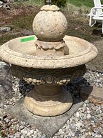 Full view of decorative concrete fountain showing the round finial and carved ornamental base, placed on gravel outdoors.
