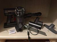 Dyson handheld vacuum with attachments arranged on a shelf, showing the main vacuum unit, charging adapter with cable, crevice tool, flat nozzle, and mini motorized brush head.