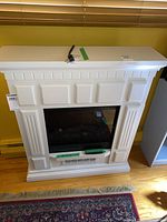 Front view of white electric fireplace with decorative mantle, power cord and remote on top.