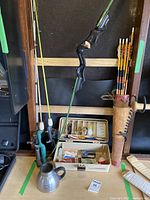 Photo showing green Prana Archery Modelo Caza recurve bow with attached string, leather quiver holding several decorated arrows, two fishing rods with handles, and open fishing tackle box showing fishing gear inside.