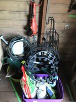 View showing electric Black & Decker hedge trimmer hanging on wall, coiled garden hose, plastic watering cans, garden fencing, black plastic garden edging, and bags of fertilizer in purple bin.