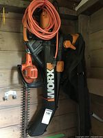 Photo of Black & Decker electric hedge trimmer hung vertically with orange extension cord wrapped at the top and WORX leaf blower with bag resting beside it.