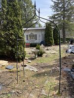 Full view of black metal garden arch standing outdoors among trees and rocks, showing overall shape and size.