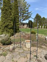 Full view of black metal garden arch standing on stone patio with greenery and trees in background