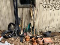 Long shot of miscellaneous garden tools leaning against a wall with clay pots in front and garden edging and tomato cages on the ground