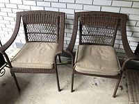 Pair of brown faux wicker patio chairs shown side by side on a concrete porch with brick wall background.