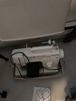 White Singer sewing machine in plastic carrying case with foot pedal and manual on floor.