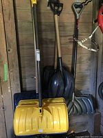 Six snow shovels leaning against a wooden wall, showing different types and colors including two yellow plastic blades and one blue blade.